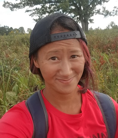 Nora Mitchell in a red shirt and backwards black baseball cap outside in a field smiling at the camera